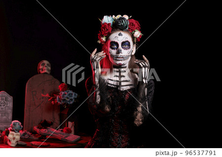 Scary goddess of death with flowers wreath posing in studio as holy santa muerte and la cavalera catrina. Young woman acting glamorous with black and white costume, day of the dead tradition. Scary goddess of death with flowers wreath posing in studio as holy santa muerte and la cavalera catrina. Young woman acting glamorous with black and white costume, day of the dead tradition. 96537791