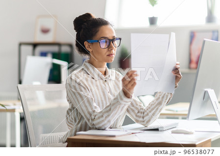 Serious busy woman in glasses sitting at table with computer and working with documents in office interior Serious busy woman in glasses sitting at table with computer and working with documents in office interior 96538077