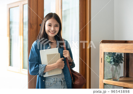 Young woman student is standing and smiling to looking on camera while holding books and bags Young woman student is standing and smiling to looking on camera while holding books and bags 96543898