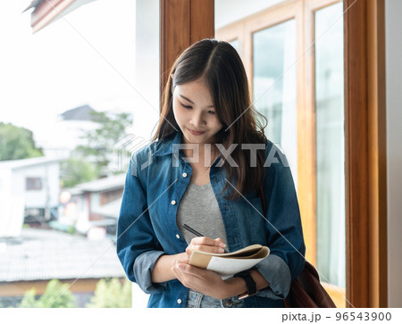 Young woman student is standing near the window to writing schedule and taking notes on notebook Young woman student is standing near the window to writing schedule and taking notes on notebook 96543900