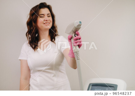 Portrait of the master of laser hair removal. A woman in a white coat holds a laser hair removal device in her hands Portrait of the master of laser hair removal. A woman in a white coat holds a laser hair removal device in her hands 96548237