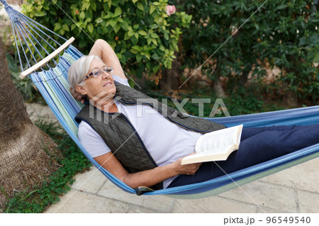 Smiling senior good-looking grey hair woman wearing glasses while reading in hammock in the summer garden 96549540