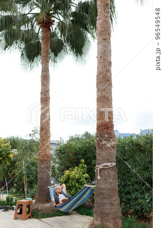 Smiling senior good-looking grey hair woman wearing glasses while reading in hammock in the summer garden under palms 96549548