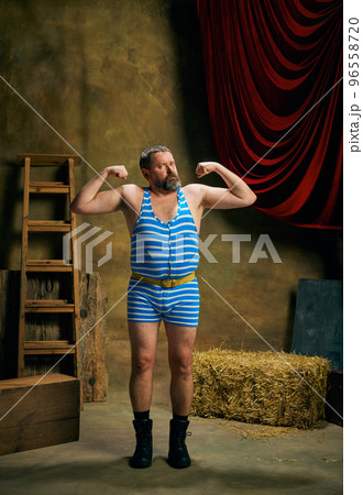 Strength. Mature man, retro circus strongman wearing striped sports swimsuit posing isolated over dark vintage circus backstage background. Art, 30s, 40s fashion style Strength. Mature man, retro circus strongman wearing striped sports swimsuit posing isolated over dark vintage circus backstage background. Art, 30s, 40s fashion style 96558720