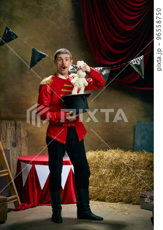 Magician. Vintage portrait of male retro circus entertainer expresses rejoice and announces start of show over dark retro circus backstage background. 96558750