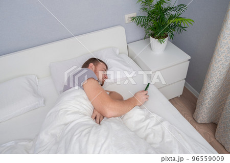 A man with a beard uses phone in his home bed A man with a beard uses phone in his home bed 96559009