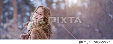 A white blonde girl in a knitted hat, a long scarf and a fur coat walks through the forest and smiles. Woman catches snow in white mittens. Winter is on the street. High quality photo. Christmas Eve 96559957