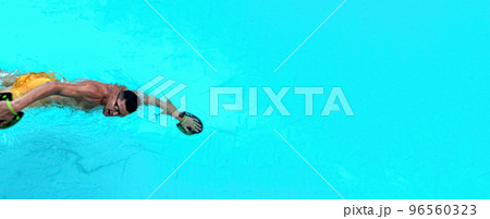 Man trains in pool. View from above on a ailetic male swimmer swimming in the blue clear water wearing goggles for swimming and using Hand Paddles. Wide banner, man at the edge of the banner 96560323