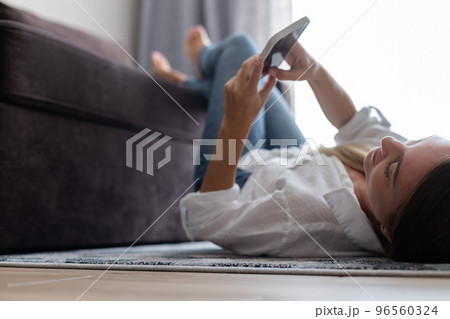 Young woman laying and relaxing on the carpet at home with mobile phone in her hands. Selective focus. 96560324
