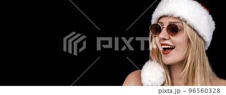 Happy Christmas blonde girl wears santa hat and sunglasses with bare shoulders looking down opening her mouth on a black background. Christmas wide banner. Holiday discounts and sales 96560328