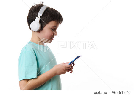 Side portrait of a concentrated teenage boy wearing wireless headphones, using mobile phone, checking social media content, surfing the Internet, browsing websites, isolated over white background. Side portrait of a concentrated teenage boy wearing wireless headphones, using mobile phone, checking social media content, surfing the Internet, browsing websites, isolated over white background. 96561570