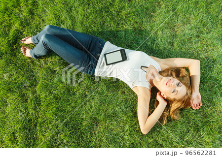 Close up portrait of an attractive young woman laying down on green grass in a park during the summer and listening to music with her earphones, being thoughtful outdoors. 96562281