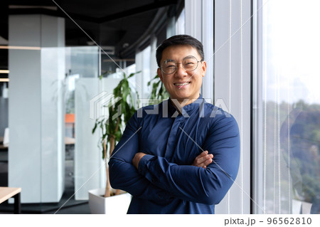Portrait of successful asian businessman, man inside office wearing shirt and glasses smiling and looking at camera, investor broker with crossed arms near window. 96562810