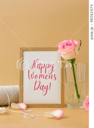Wooden frame with greeting card text HAPPY WOMENS DAY delicate pink roses, spool of white cotton rope on beige background. Minimal trendy composition. Romantic pastel pink rose flowers. Neutral earth 96562974