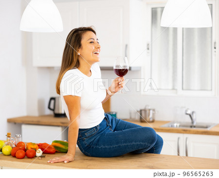 Smiling young Hispanic woman drinking wine sitting on table in kitchen 96565263