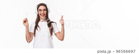 Waist-up portrait of excited, smiling happy and enthusiastic young girl in headphones, holding wire of earphones and show thumbs-up, ready to plug in to smartphone and listen some music Waist-up portrait of excited, smiling happy and enthusiastic young girl in headphones, holding wire of earphones and show thumbs-up, ready to plug in to smartphone and listen some music 96566697