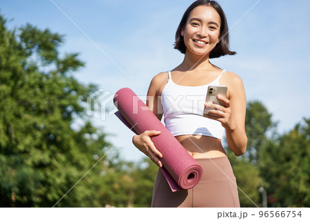 Portrait of young asian woman checking her phone during workout, walking in park with rubber mat, going to the gym, holding smartphone Portrait of young asian woman checking her phone during workout, walking in park with rubber mat, going to the gym, holding smartphone 96566754