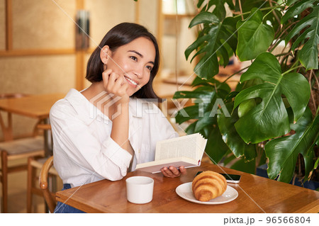 Dreamy young smiling asian woman reading book, sitting in cafe, eating croissant and drinking coffee in cozy interior Dreamy young smiling asian woman reading book, sitting in cafe, eating croissant and drinking coffee in cozy interior 96566804