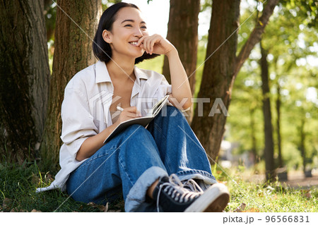 Portrait of young asian woman writing in her notebook, expressing her thoughts on paper in diary, smiling and sitting under tree on summer day in park Portrait of young asian woman writing in her notebook, expressing her thoughts on paper in diary, smiling and sitting under tree on summer day in park 96566831