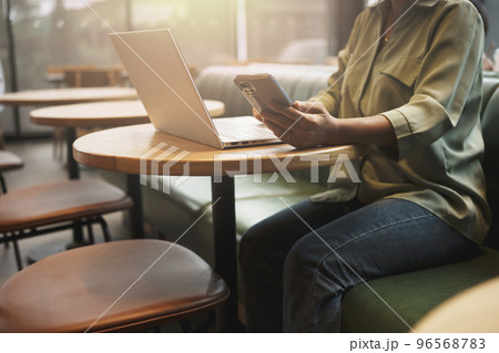 Close-up of woman's hand using smartphone while working on laptop in a coffee shop. Close-up of woman's hand using smartphone while working on laptop in a coffee shop. 96568783