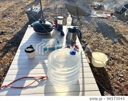 焚き火とアウトドアテーブルに乗せたウォータータンクと鍋など 96573043