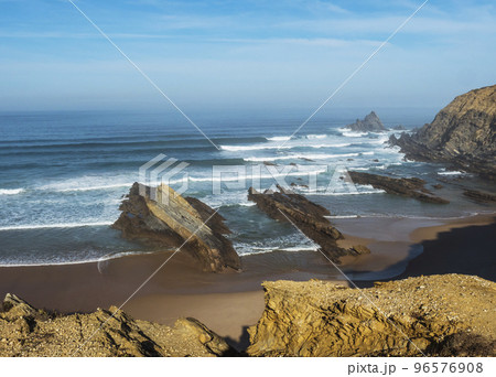 View of empty Alteirinhos sand beach with ocean waves, cliffs, stones and rocks in morning golden light at wild Rota Vicentina coast, Odemira, Portugal. 96576908