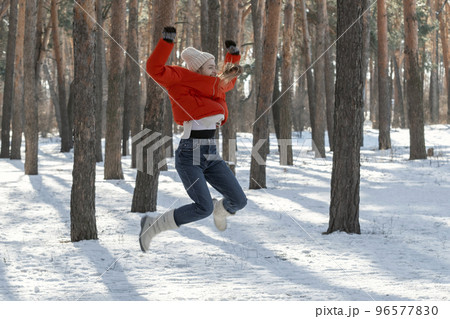 Cheerful young woman jumps high in snowy forest. Walk through the winter wood on sunny day 96577830
