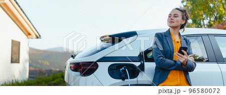Young woman with smartphone waiting while her electric car charging in home charging station, sustainable and economic transportation concept. 96580072