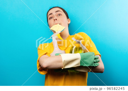 Portrait of a surprised caucasian young woman in rubber gloves, holding a sponge in her mouth and household cleaning products in her hands. Blue background. Cleaning day concept Portrait of a surprised caucasian young woman in rubber gloves, holding a sponge in her mouth and household cleaning products in her hands. Blue background. Cleaning day concept 96582245
