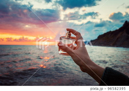 Female hand spraying a golden transparent perfume bottle. Close-up. In the background, the ocean and cloudy sunset. Concept of perfume advertising 96582395