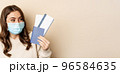 Travel and covid-19 pandemic. Close up portrait of smiling woman in medical mask, showing passport with two tickets, concept of tourism, beige background 96584635