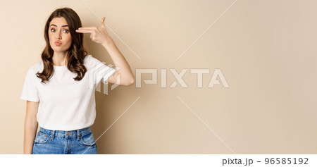 Young woman showing finger pistol sign near head and looking annoyed, sick and tired, standing over beige background Young woman showing finger pistol sign near head and looking annoyed, sick and tired, standing over beige background 96585192