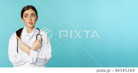 Portrait of indecisive, confused female doctor, pharmacy worker pointing sideways and biting lip clueless, dont know, standing over blue background 96585269