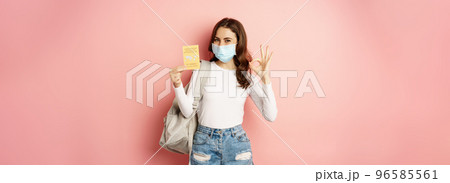 Young woman in medical mask, showing okay sign and covid vaccination certificate, travel during pandemic, going on holiday, standing over pink background 96585561