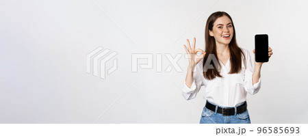 Happy smiling woman with satisfied face, recommending excellent app, showing mobile phone screen, okay sign, standing over white background 96585693