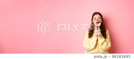 Image of coy attractive girl thinking of something interesting, showing her smile with magnifying glass near lips, standing against pink background, looking aside at promotional text 96585695