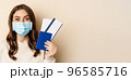 Travel and covid-19 pandemic. Close up portrait of smiling woman in medical mask, showing passport with two tickets, concept of tourism, beige background 96585716