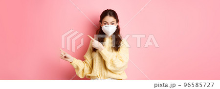 Covid, corona and social distancing concept. Excited female model in respirator shows you advertisement, points at logo aside, wears medical respirator on face, pink background Covid, corona and social distancing concept. Excited female model in respirator shows you advertisement, points at logo aside, wears medical respirator on face, pink background 96585727