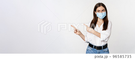 Portrait of corporate woman in face medical mask, pointing fingers left, showing logo, company banner, standing against white background Portrait of corporate woman in face medical mask, pointing fingers left, showing logo, company banner, standing against white background 96585735