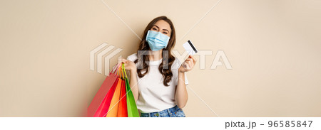 Covid-19 pandemic and lifestyle concept. Young woman posing in medical face mask with shopping bags from mall, holding credt card in hand, beige background 96585847