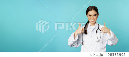 Happy woman doctor, medical worker in white coat, showing thumbs up in approval, like something, praise, standing over blue background Happy woman doctor, medical worker in white coat, showing thumbs up in approval, like something, praise, standing over blue background 96585995