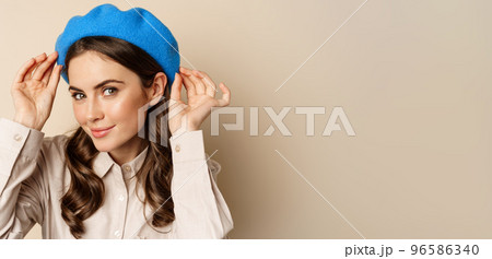 Stylish modern girl put on trendy hat on head and smiling, going out, posing against beige background 96586340