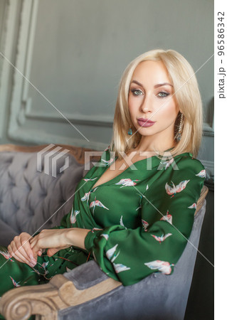 Beautiful blond woman with makeup in green dress sitting on grey velvet sofa Beautiful blond woman with makeup in green dress sitting on grey velvet sofa 96586342