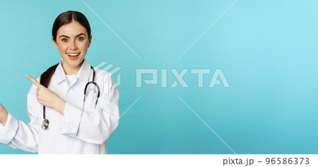 Portrait of smiling medical worker, girl doctor in white coat with stethoscope, pointing fingers left, showing medical clinic advertisement, torquoise background 96586373