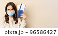Travel and covid-19 pandemic. Girl in medical face mask travelling, showing passport with two tickets on plane, standing against beige background 96586427
