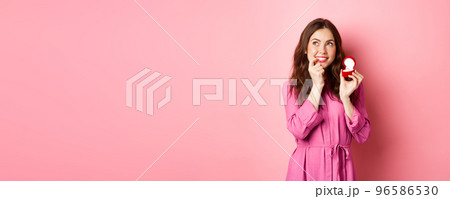 Beautiful girl dream of her wedding day, looking thoughtful up, showing engagement ring in red box and smiling, standing over pink background Beautiful girl dream of her wedding day, looking thoughtful up, showing engagement ring in red box and smiling, standing over pink background 96586530