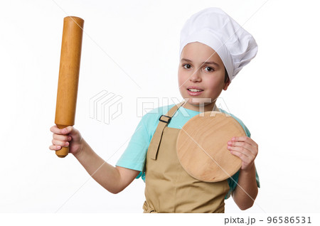 Adorable Caucasian child boy, wearing a chef's hat and beige apron, posing with a rolling pin and wooden cutting board, isolated on white background. Baker Confectioner. Chef pastry. Cook profession Adorable Caucasian child boy, wearing a chef's hat and beige apron, posing with a rolling pin and wooden cutting board, isolated on white background. Baker Confectioner. Chef pastry. Cook profession 96586531