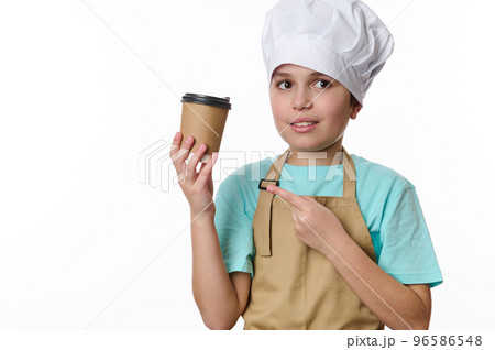 Hispanic handsome preteen boy dressed as bartender barista in beige chef apron and hat, points at a takeaway hot coffee tea drink in eco paper cup, isolated on white background. Free advertising space 96586548