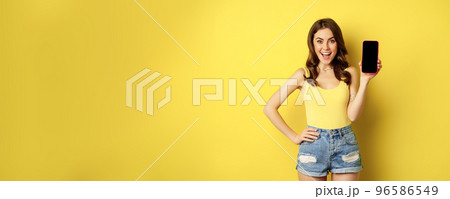 Stylish smiling woman showing mobile phone screen, app interface on smartphone, standing in tank top and shorts against yellow background 96586549