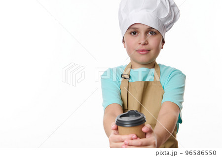 Cheerful teenage boy wearing a white chef's hat and beige apron, barista bartender handing hot coffee drink in a paper cup to go, isolated over white background. Free space for your advertising text 96586550